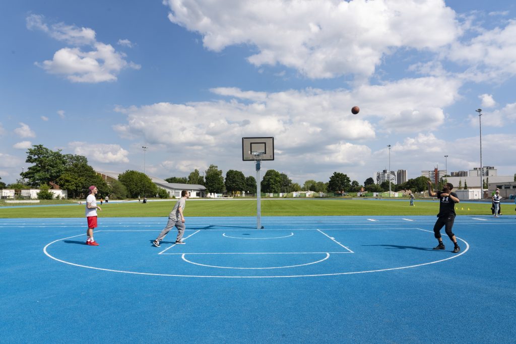 Auf einem der Franklin Sportanlage mit blauen Sportplatz spielen drei Männer Basketball. Der Ball fliegt durch die Luft.